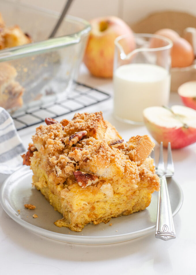 A slice of apple French toast casserole on a grey plate with apples in the background.