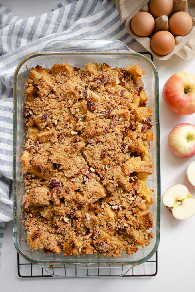 A pan of apple French toast casserole on a wire cooling rack with a striped linen tea towel next to it, along with some eggs and apples.