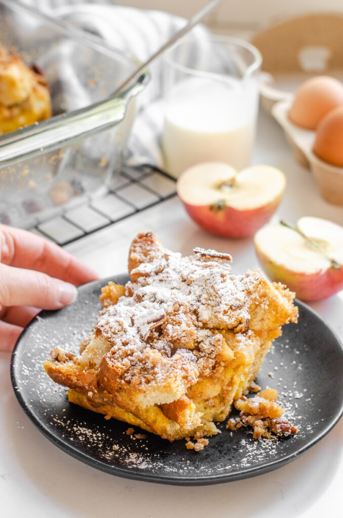 A slice of apple French toast casserole with apples in the background and powdered sugar on top.