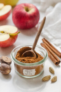 A small jar of homemade apple pie spice blend with spices, apples, and a linen napkin around it, and a mini wooden spoon in the jar.