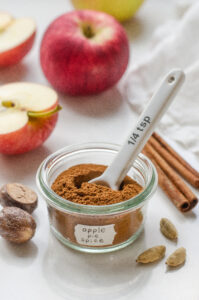 A small jar of homemade apple pie spice blend with spices, apples, and a linen napkin around it, and a small measuring spoon inside it.