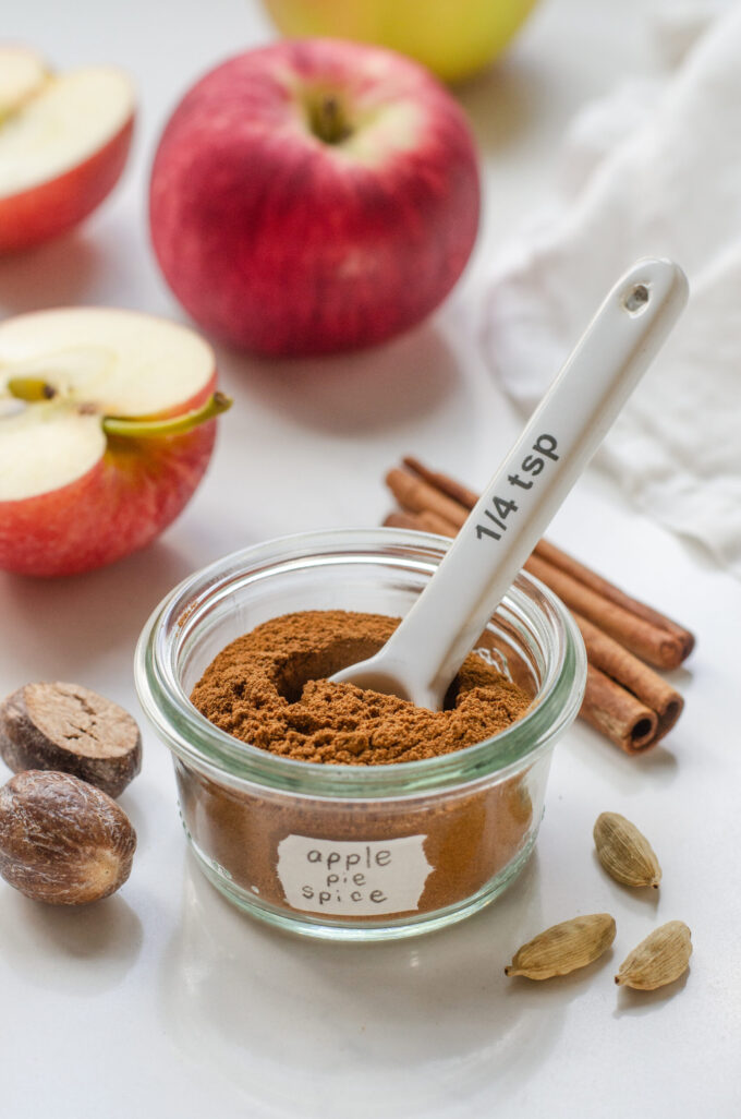 A small jar of homemade apple pie spice blend with spices, apples, and a linen napkin around it, and a small measuring spoon inside it.