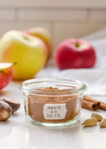 A small jar of homemade apple pie spice blend with spices, apples, and a linen napkin around it.