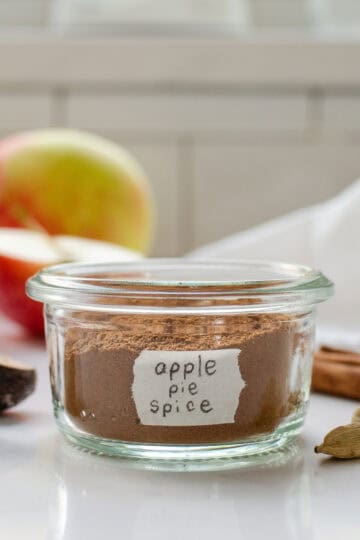 A small jar of homemade apple pie spice blend with spices, apples, and a linen napkin around it.