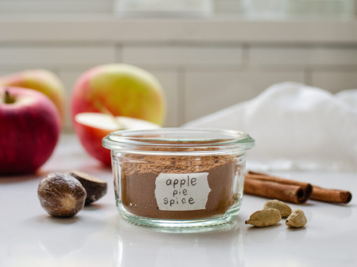 A small jar of homemade apple pie spice blend with spices, apples, and a linen napkin around it.