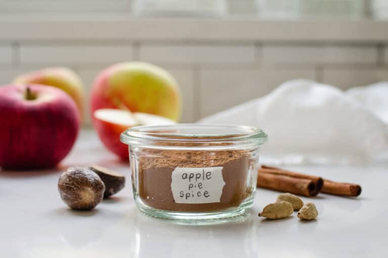 A small jar of homemade apple pie spice blend with spices, apples, and a linen napkin around it.