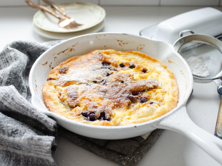 A skillet of Sourdough Dutch Baby with a linen tea towel to one side, and a butter dish and sifter to the other side.