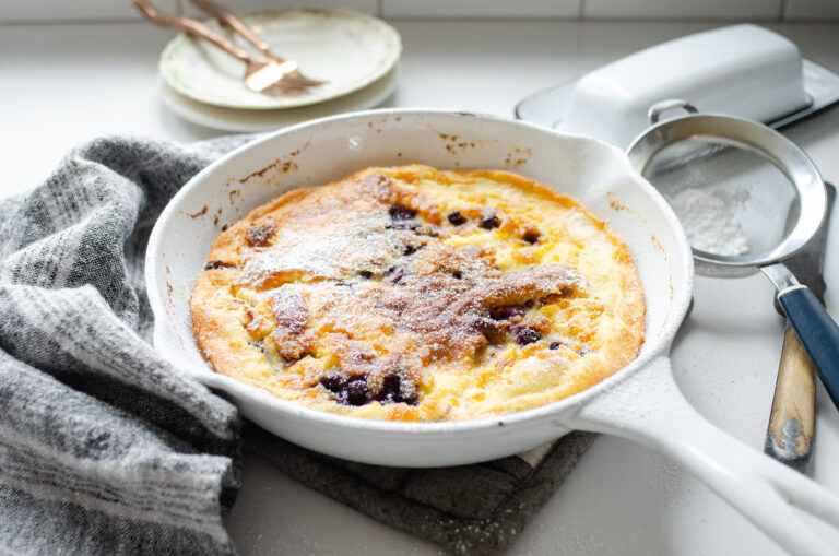 A skillet of Sourdough Dutch Baby with a linen tea towel to one side, and a butter dish and sifter to the other side.