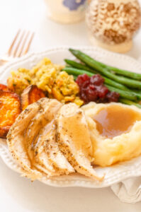 A plate of Thanksgiving dinner featuring green beans, mashed potatoes, gravy, stuffing, sweet potatoes, cranberry sauce, and chicken breast.