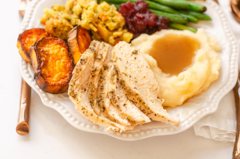 A plate of Thanksgiving dinner featuring green beans, mashed potatoes, gravy, stuffing, sweet potatoes, cranberry sauce, and chicken breast.