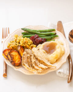 A plate of Thanksgiving dinner featuring green beans, mashed potatoes, gravy, stuffing, sweet potatoes, cranberry sauce, and chicken breast.