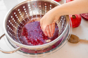 A bowl of water with a colander inside and pomegranate arils in it.