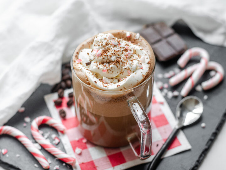 A mug of Peppermint Mocha (a Starbucks recipe) with a red checked napkin underneath and candy canes, coffee beans, and chocolate in the background.