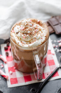 A mug of Peppermint Mocha (a Starbucks recipe) with a red checked napkin underneath and candy canes, coffee beans, and chocolate in the background.