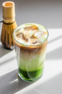 A 12 ounce glass of matcha coffee on a sunny white countertop with a matcha whisk in the background and a white backsplash in the background