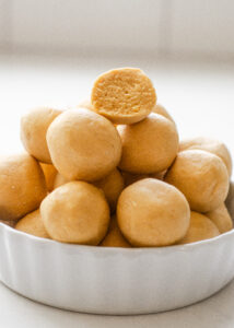 A ramekin filled with no bake powdered sugar peanut butter balls (without chocolate) with one on top cut in half so you can see the inside.