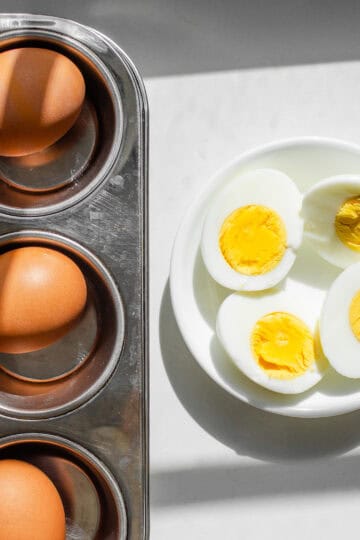 A muffin tin with hard boiled eggs inside with two peeled and halved eggs in a bowl next to it.