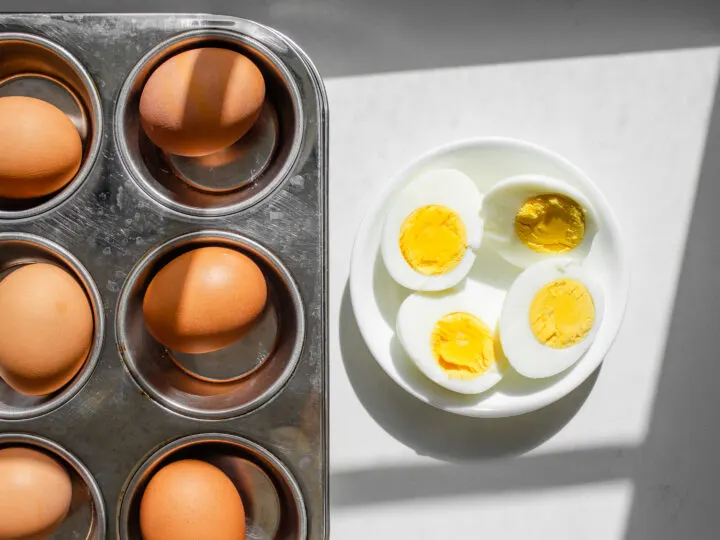 A muffin tin with hard boiled eggs inside with two peeled and halved eggs in a bowl next to it.