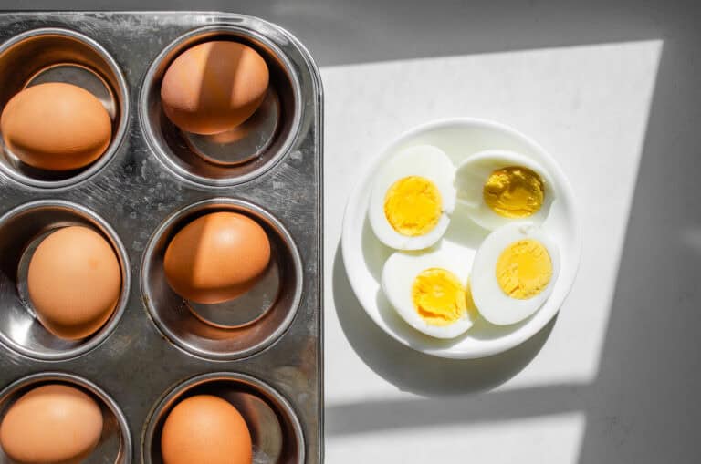 A muffin tin with hard boiled eggs inside with two peeled and halved eggs in a bowl next to it.