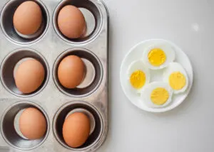 A muffin tin with hard boiled eggs inside with two peeled and halved eggs in a bowl next to it.