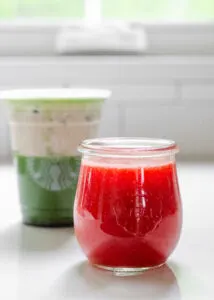 A jar of homemade Starbucks strawberry puree sauce with an iced matcha latte with strawberry cold foam on top in the background.