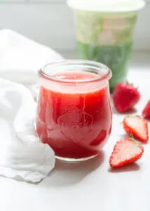 A jar of homemade Starbucks strawberry puree on a white countertop with fresh strawberries beside it.