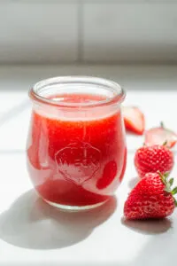 A jar of homemade Starbucks strawberry puree on a white countertop with fresh strawberries beside it.