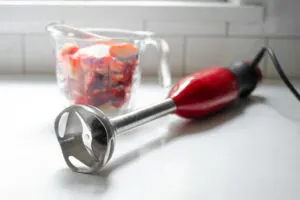 A stick blender on a white countertop with fresh strawberries in a measuring cup behind it.