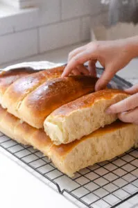 A stack of homemade sourdough hotdog buns on a black wire cooling rack with a linen napkin off to the side.