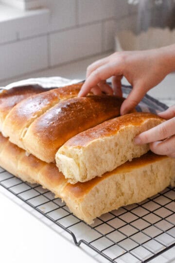 A stack of homemade sourdough hotdog buns on a black wire cooling rack with a linen napkin off to the side.