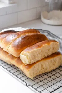 A stack of homemade sourdough hotdog buns on a black wire cooling rack with a linen napkin off to the side.