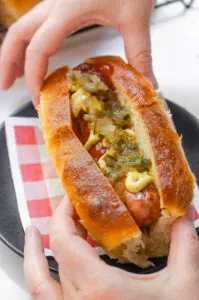 A sourdough hotdog bun with a brat inside with ketchup, mustard, and relish on top.