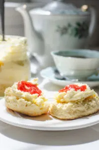 A scone spread with clotted cream with a dab of jam on top and a teacup and teapot behind it.