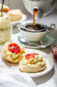 A scone spread with clotted cream with a dab of jam on top and a teacup and teapot behind it.