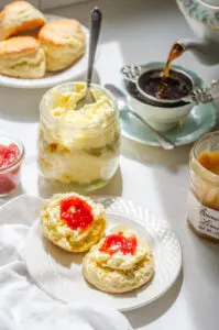 A scone spread with clotted cream with a dab of jam on top and a teacup and teapot behind it.