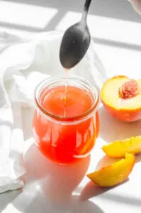 Peach syrup in a jar with fresh peaches and a white napkin around it.