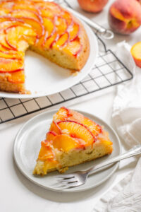 A slice of peach olive oil cake on a grey p late with a fork on it and the rest of the cake in the background.