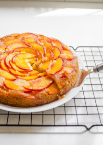 A peach olive oil cake on a plate on a wire rack with a slice cut.