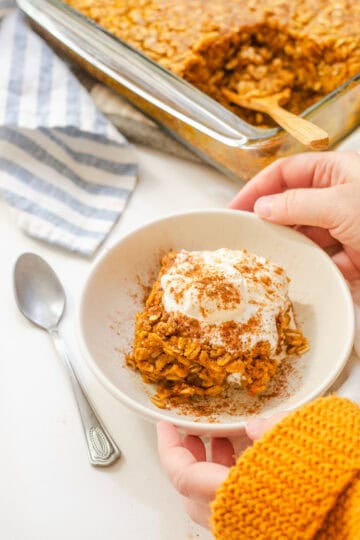 A bowl of pumpkin pie baked oatmeal with a dollop of maple whipped cream on top and the pan in the background and hands holding the bowl.