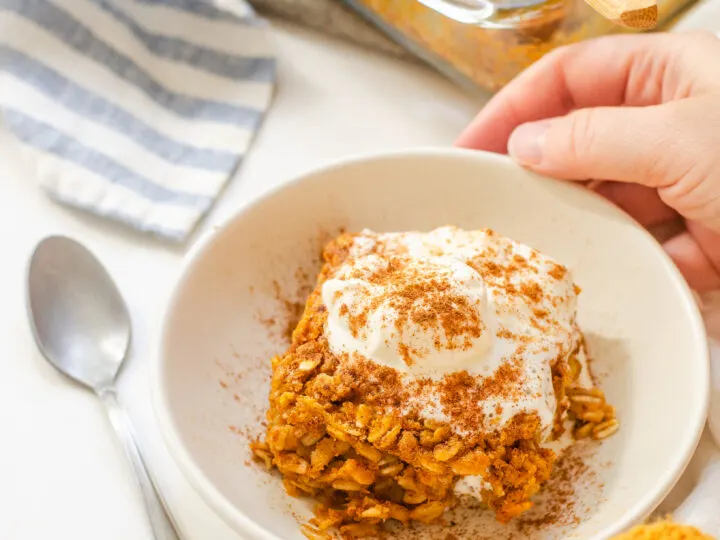 A bowl of pumpkin pie baked oatmeal with a dollop of maple whipped cream on top and the pan in the background and hands holding the bowl.