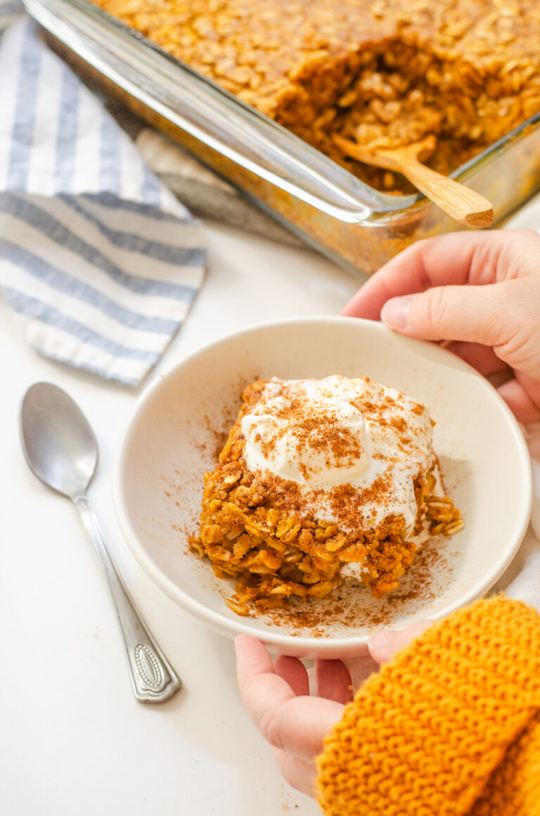 A bowl of pumpkin pie baked oatmeal with a dollop of maple whipped cream on top and the pan in the background and hands holding the bowl.