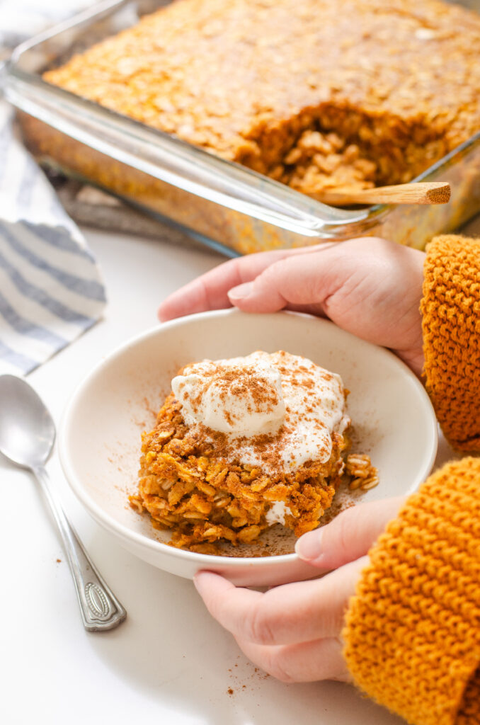 A bowl of pumpkin pie baked oatmeal with a dollop of maple whipped cream on top and the pan in the background and hands holding the bowl.