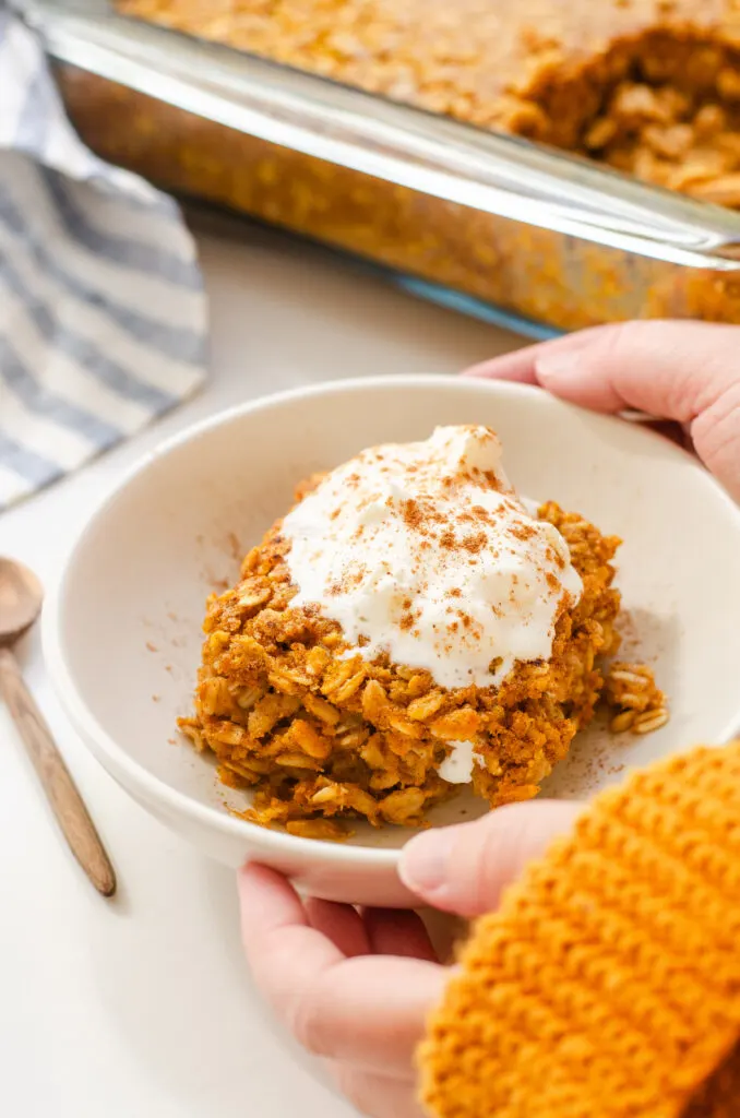 A bowl of pumpkin pie baked oatmeal with a dollop of maple whipped cream on top and the pan in the background and hands holding the bowl.