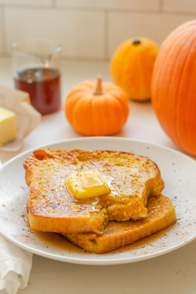 Two slices of pumpkin French toast on a plate with butter and maple syrup on top, and pumpkins in the background.