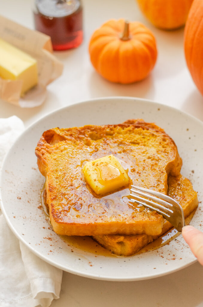 Two slices of pumpkin French toast on a plate with butter and maple syrup on top, and pumpkins in the background.