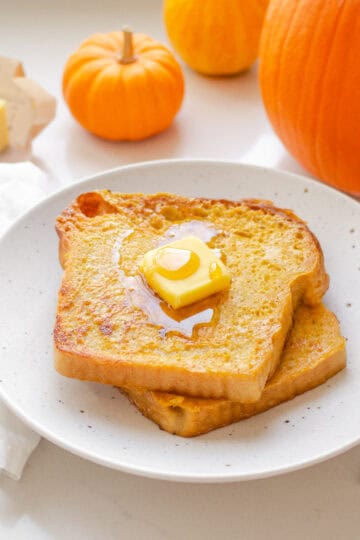 Two slices of pumpkin French toast on a plate with butter and maple syrup on top, and pumpkins in the background.
