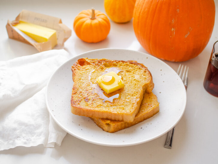 Two slices of pumpkin French toast on a plate with butter and maple syrup on top, and pumpkins in the background.