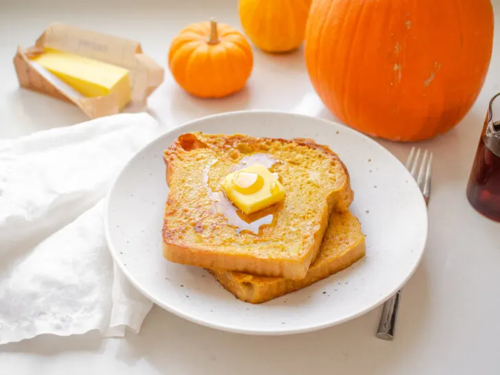 Two slices of pumpkin French toast on a plate with butter and maple syrup on top, and pumpkins in the background.
