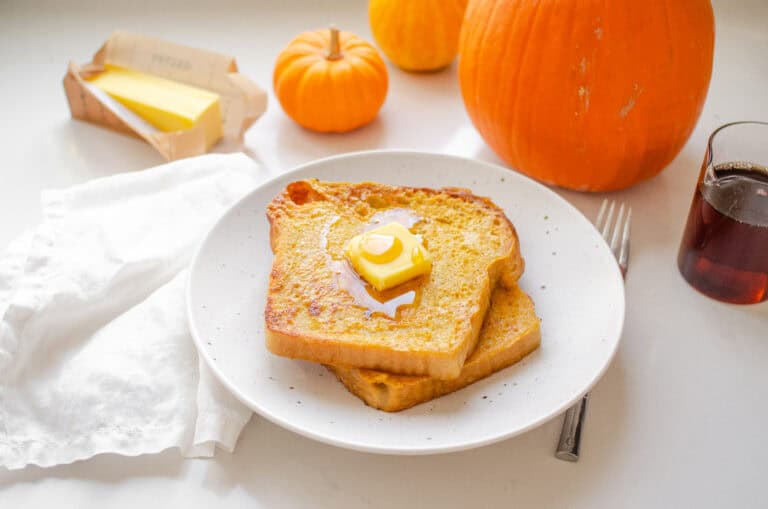 Two slices of pumpkin French toast on a plate with butter and maple syrup on top, and pumpkins in the background.
