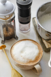 A latte made with a French press and hand held frother on a white countertop with the French press behind the latte mug.